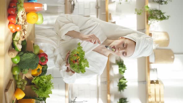 Happy Woman in a White Coat and with a Towel on Her Head Eats a Salad of Fresh Vegetables at Home