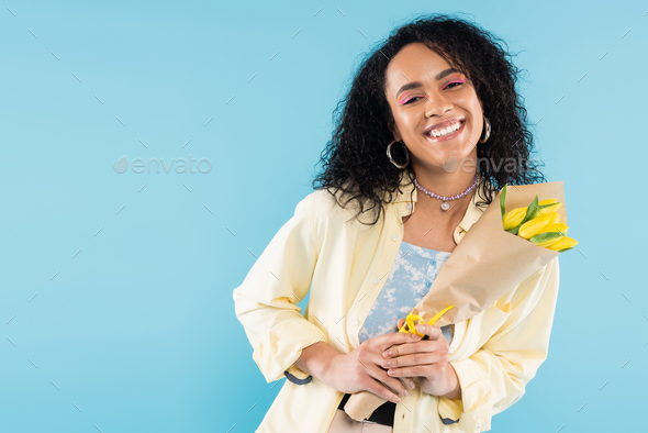trendy african american woman in hoop earrings and beads holding yellow tulips and smiling at camera