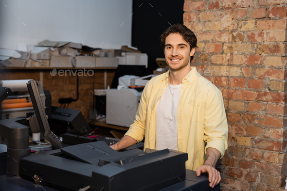 happy man in yellow shirt standing near professional equipment in print center