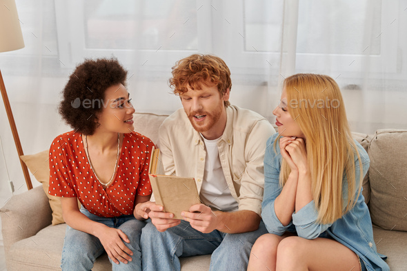open relationships, redhead man reading book to multicultural women in living room, non traditional