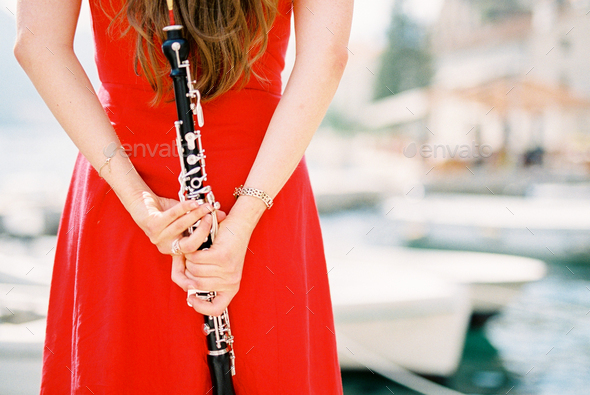 Girl-musician with a clarinet in her hands behind her back stands at the pier. Cropped. Back view