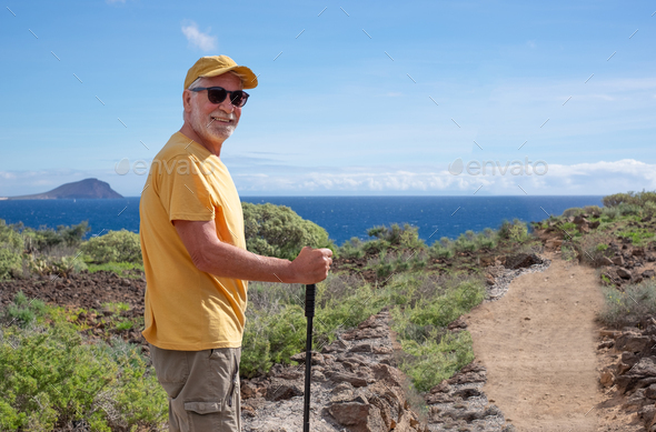 Happy senior man in yellow cap and t-shirt outdoors on hike in countryside with sea view.