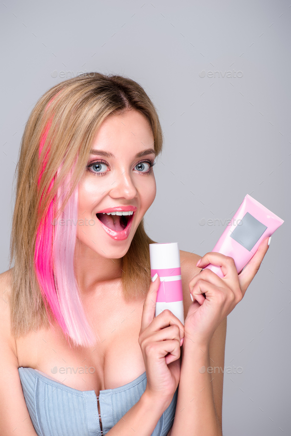 excited young woman with colored bob cut holding hair care supplies and looking at camera isolated