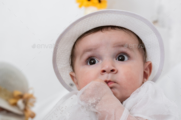 six months old baby girl with a very curious look raised to the sky, dressed in a white hat.