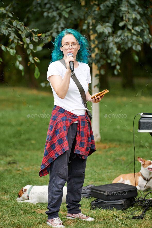 Young adult woman host of event in round glasses with turquoise dyed hair speaks into microphone