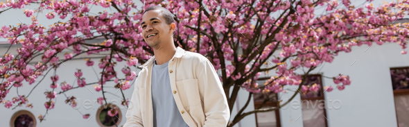 positive african american man in shirt jacket walking near cherry tree, banner