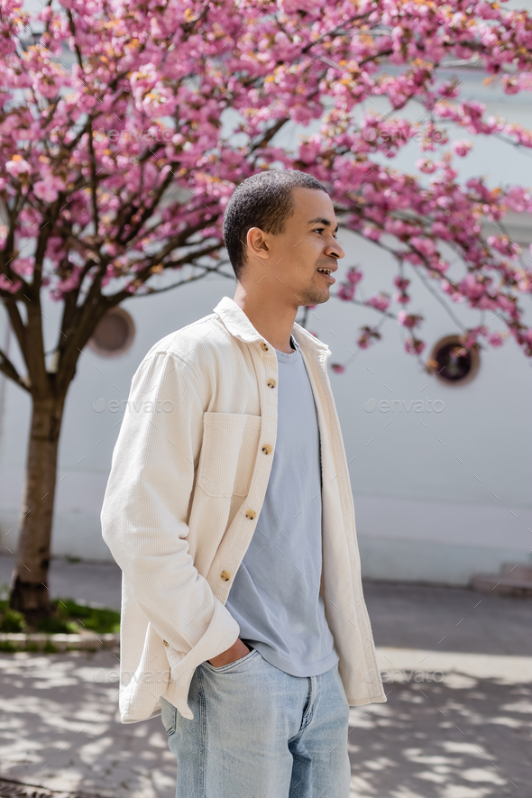 young african american man in shirt jacket walking with hand in pocket near cherry tree