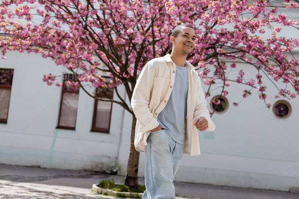 positive african american man in shirt jacket walking with hand in pocket near cherry tree