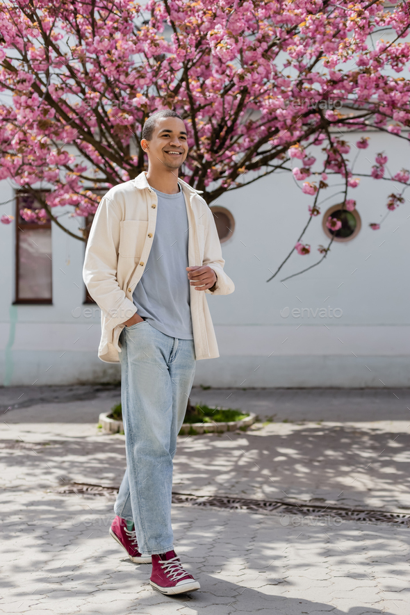 happy young african american man in shirt jacket walking with hand in pocket near cherry tree