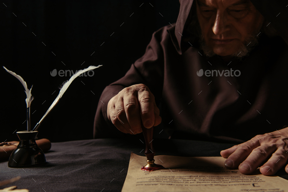 medieval monk in dark robe with hood stamping parchment with wax seal isolated on black