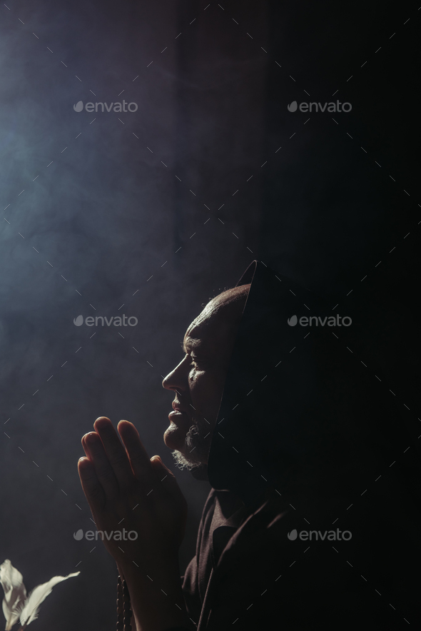 side view of senior monk in hood praying at night on black background with copy space