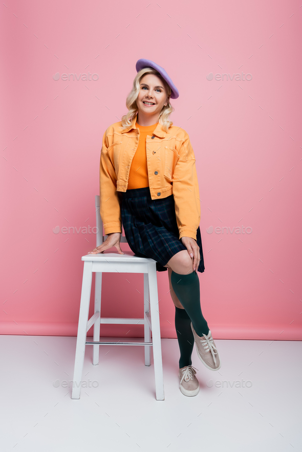 Smiling woman in skirt and beret posing near chair on pink background