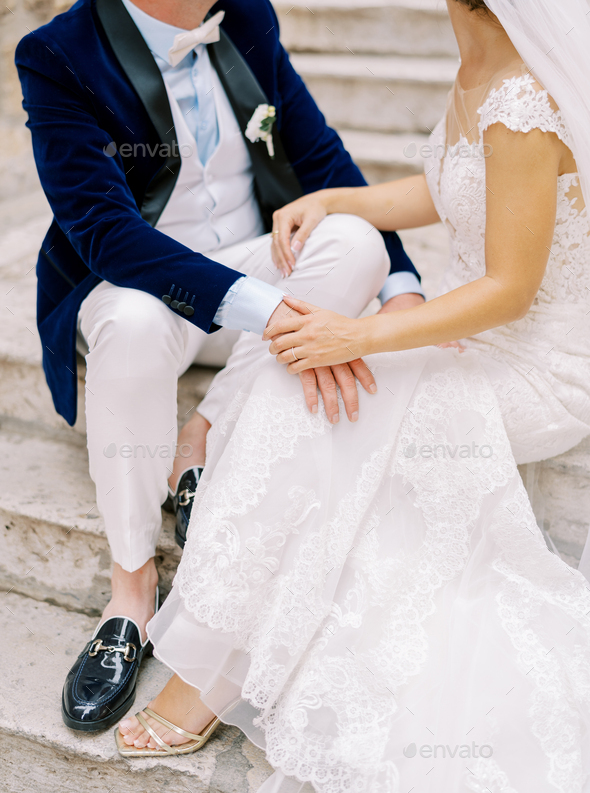 Bride and groom sit on old stone steps and place their hands on each other laps. Cropped. Faceless