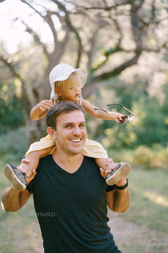 Little girl holds her dad sunglasses in her hand while sitting on his  shoulders Stock Photo by Nadtochii