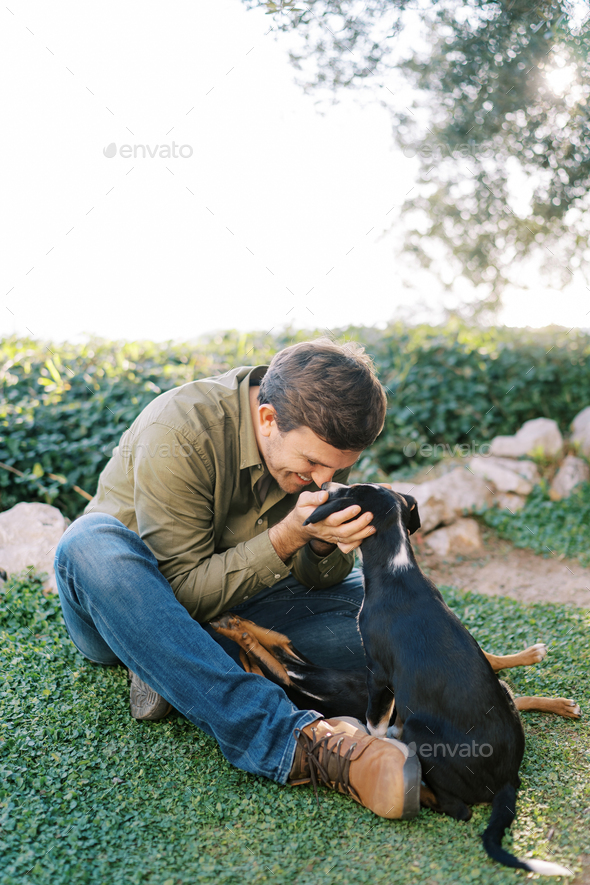 Smiling young man sits on the lawn and touches with his nose the nose of a big puppy