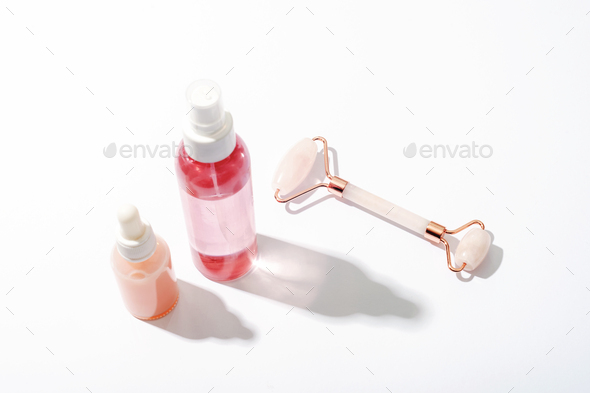 Pink cosmetic bottles and rose quartz crystal facial roller on white background with strong shadows