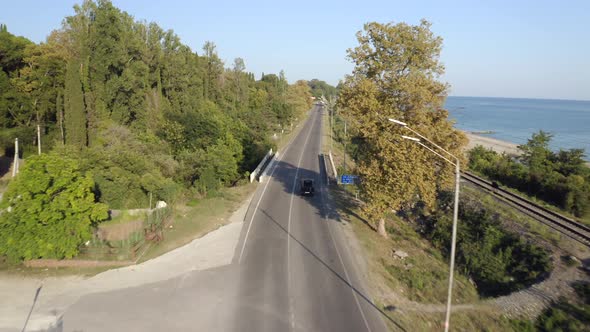 Black Suv Car Drives Along Grey Asphalt Coastal Road Between Green Forest alt