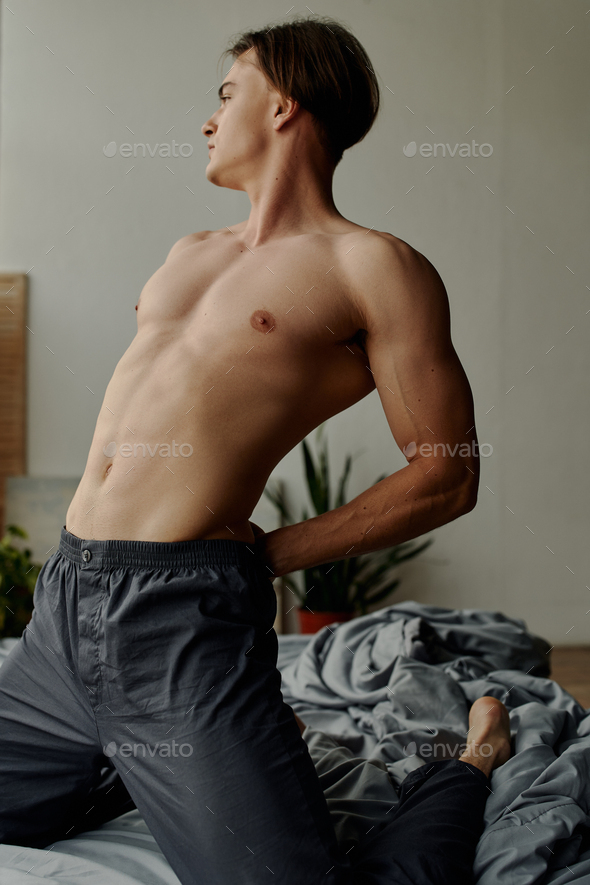 sexy shirtless man in pajama pants stretching back while kneeling on bed