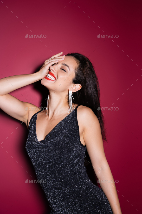 elegant woman in shiny earrings and black lurex dress obscuring face and laughing on red background