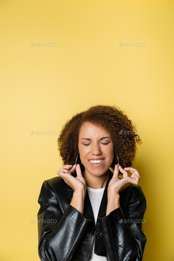 pleased african american woman in stylish leather jacket touching hoop earrings on yellow