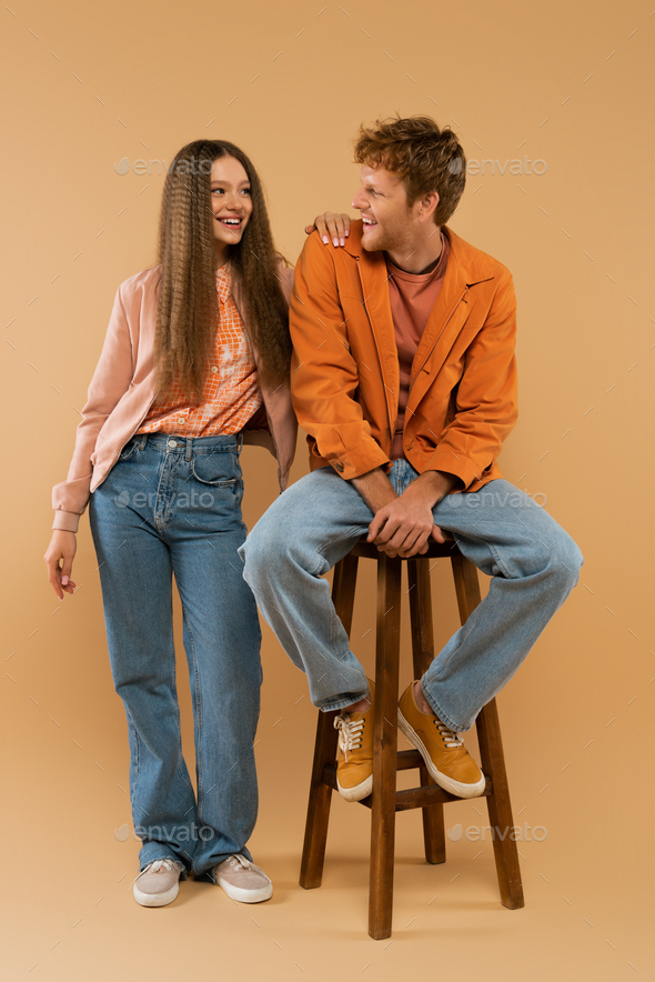 full length of young redhead man sitting on high chair and looking at girlfriend with wavy hair on