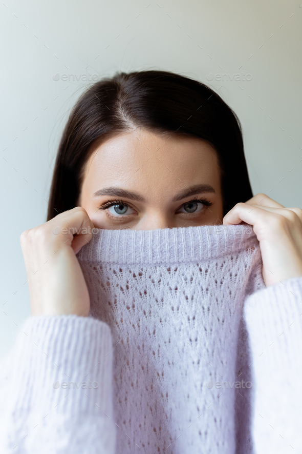 brunette woman obscuring face with collar of warm knitted sweater while looking at camera isolated