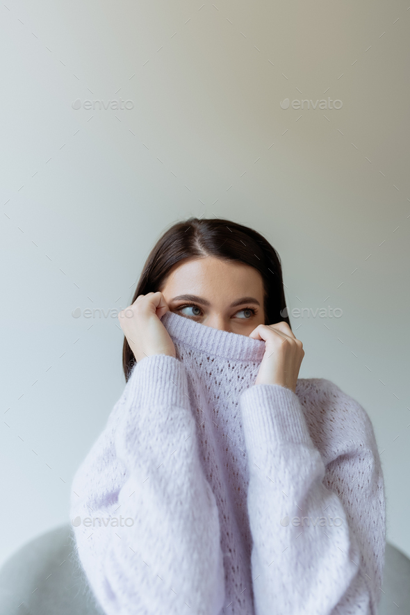young brunette woman obscuring face with warm knitted sweater and looking away isolated on grey