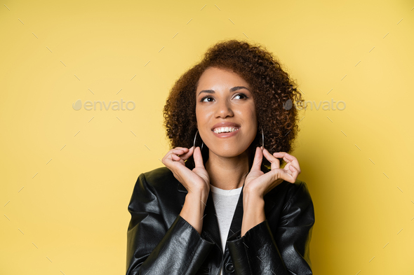 happy african american woman in stylish leather jacket touching hoop earrings on yellow