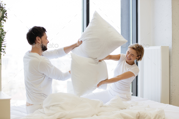 Side view of happy Caucasian husband and wife having pillow fight in bed