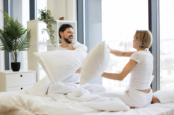 Side view of happy Caucasian husband and wife having pillow fight in bed
