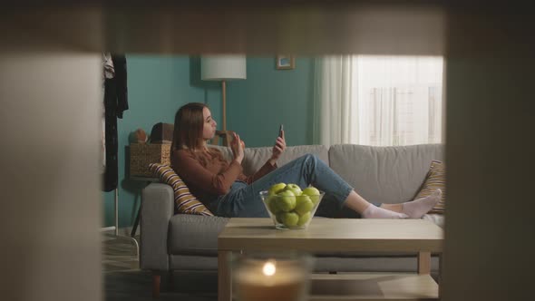 Young Girl Eats a Red Apple Lying on Sofa and and Speaks Via Video Link alt
