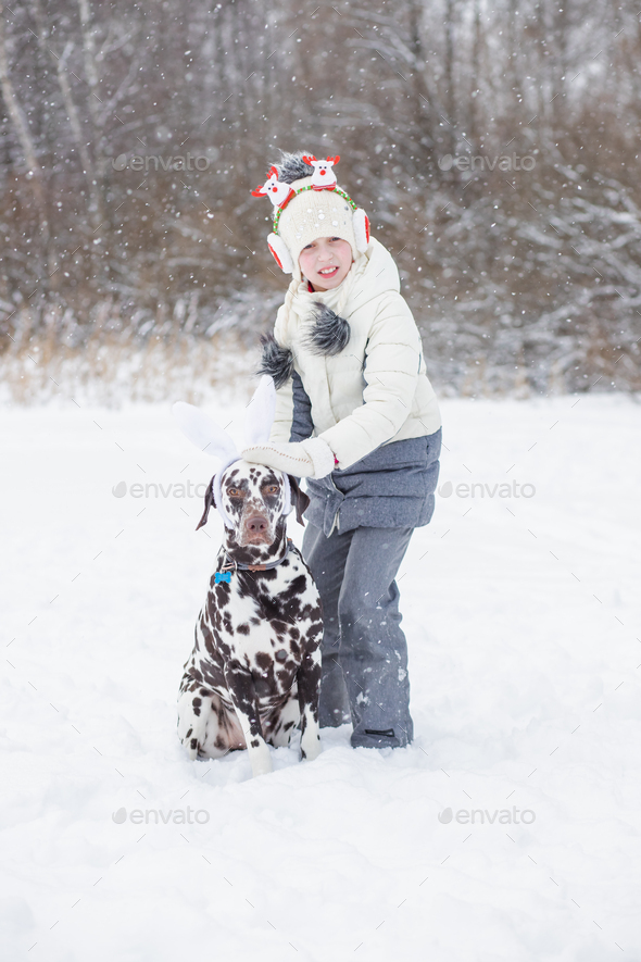 Girl wearing winter warm coat, hat and Christmas headband has fun with dalmatian dog outdoor. new