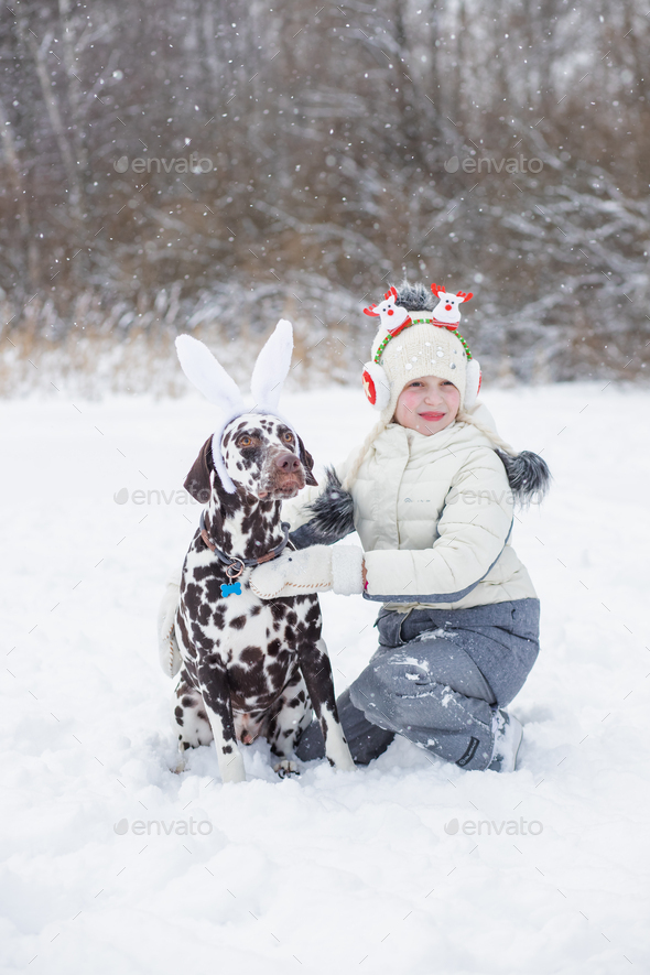 Girl wearing winter warm coat, hat and Christmas headband has fun with dalmatian dog outdoor. new
