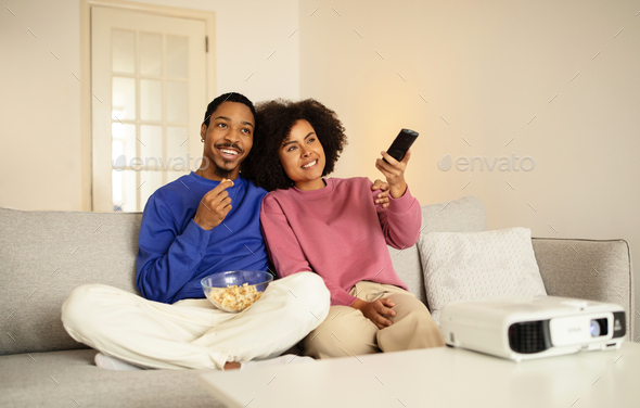 Smiling African spouses watching film on TV, munching popcorn indoor
