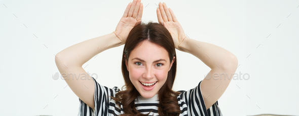 Image of cute brunette girl shows animal eats with hands on top of head, smiling and looking happy