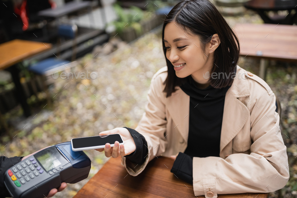 Smiling asian woman in trench coat paying with smartphone near waiter holding payment terminal on