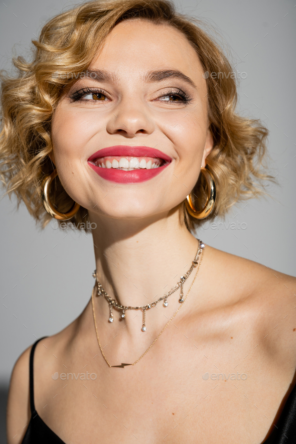 portrait of happy young woman in hoop earrings and silver necklace smiling isolated on grey
