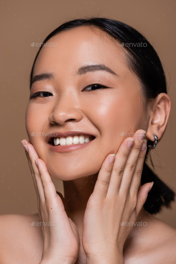 Portrait Of Babe Asian Woman With Naked Shoulders Touching Cheeks Isolated On Brown Stock Photo