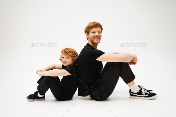 full length of happy red haired dad and son in black t-shirts and pants sitting back to back on