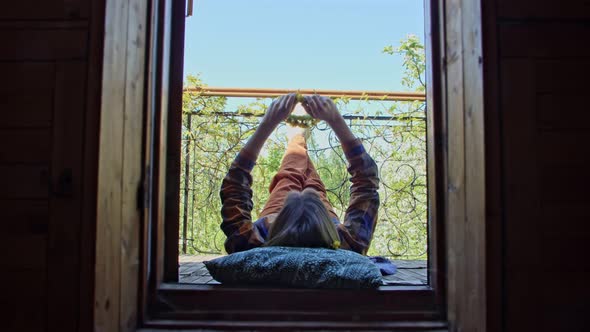 the Woman on the Balcony Lies Freely Raising Her Legs Up and Weaving a Wreath of Flowers Enjoying alt