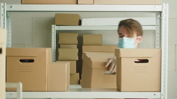 Man Wearing Medical Face Mask and Protective Gloves Putting Parcel on Shelf Employee of Warehouse alt