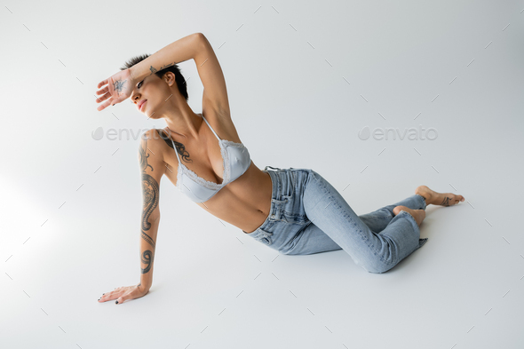 full length of sexy barefoot woman in silk bralette and blue jeans obscuring face with hand while