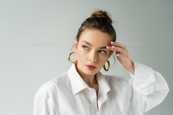 portrait of young woman in white shirt and hoop earrings touching eyebrow and looking away isolated