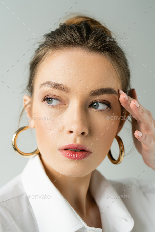 portrait of young woman with natural makeup and hoop earrings touching face and looking away