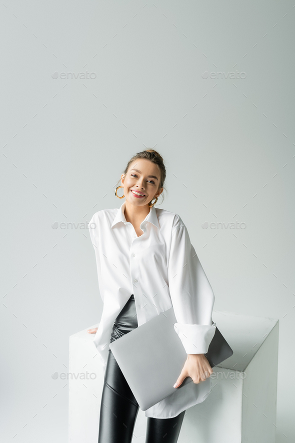 joyful woman in hoop earrings and oversize shirt holding laptop and looking at camera near white