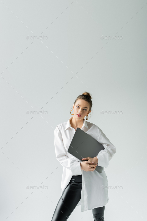 trendy woman in white shirt and hoop earrings posing with laptop and looking at camera isolated on