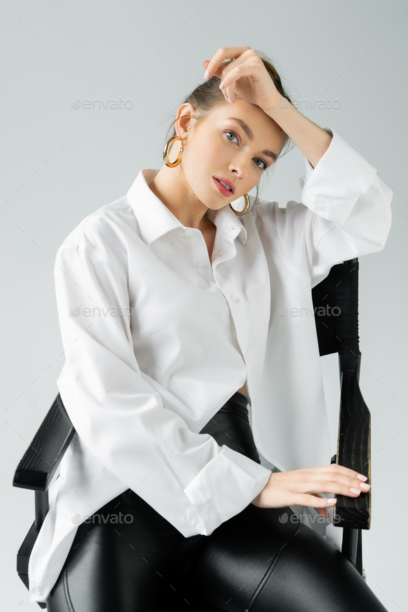 stylish woman in hoop earrings and white oversize shirt posing on chair and looking at camera