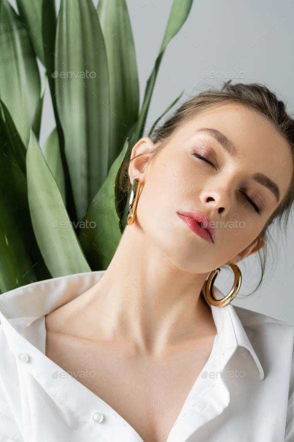 portrait of sensual woman in white shirt and hoop earrings posing with closed eyes near blurred