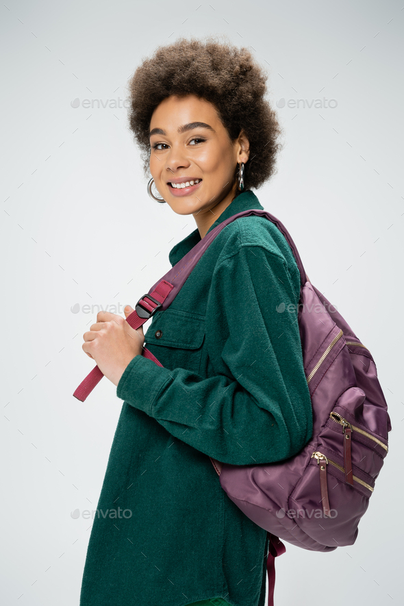 cheerful african american woman in shirt and hoop earrings holding backpack and looking at camera