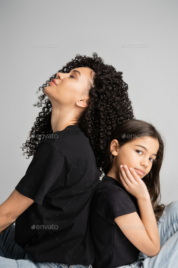 Curly mother and daughter in black t-shirts sitting back to back isolated on grey